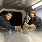 Two young men in dark clothing lean over a bench and turn to face the camera. Between them is a black fin-like object. 