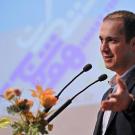Man in suit speaking at podium with microphones, gesturing near a floral arrangement