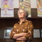 Woman in patterned blouse standing with arms crossed beneath hanging colorful art, photograph