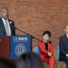 Standing at a lectern, UC Davis Chancellor Gary S. May speaks into a microphone as a seated man and woman look on