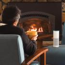 A man sits on a chair in front of a fireplace, back to the camera. A chair faces him with an Amazon Echo device sitting on it, lit up.