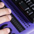 A hand rests on a purple Braille display next to a black keyboard on a wooden surface.