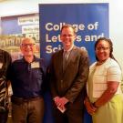 Five people stand smiling in front of a "College of Letters and Science" banner. From left: a tall man in a patterned shirt, an older man in a blue polo, a man in a suit and tie, a Black woman in a white top, and an older man in a yellow shirt. Colorful balloons are visible in the background.