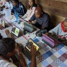 Farmers in Malagasy sit on each side of a rectangular table lined with a quilted tablecloth playing a game on digital tablets